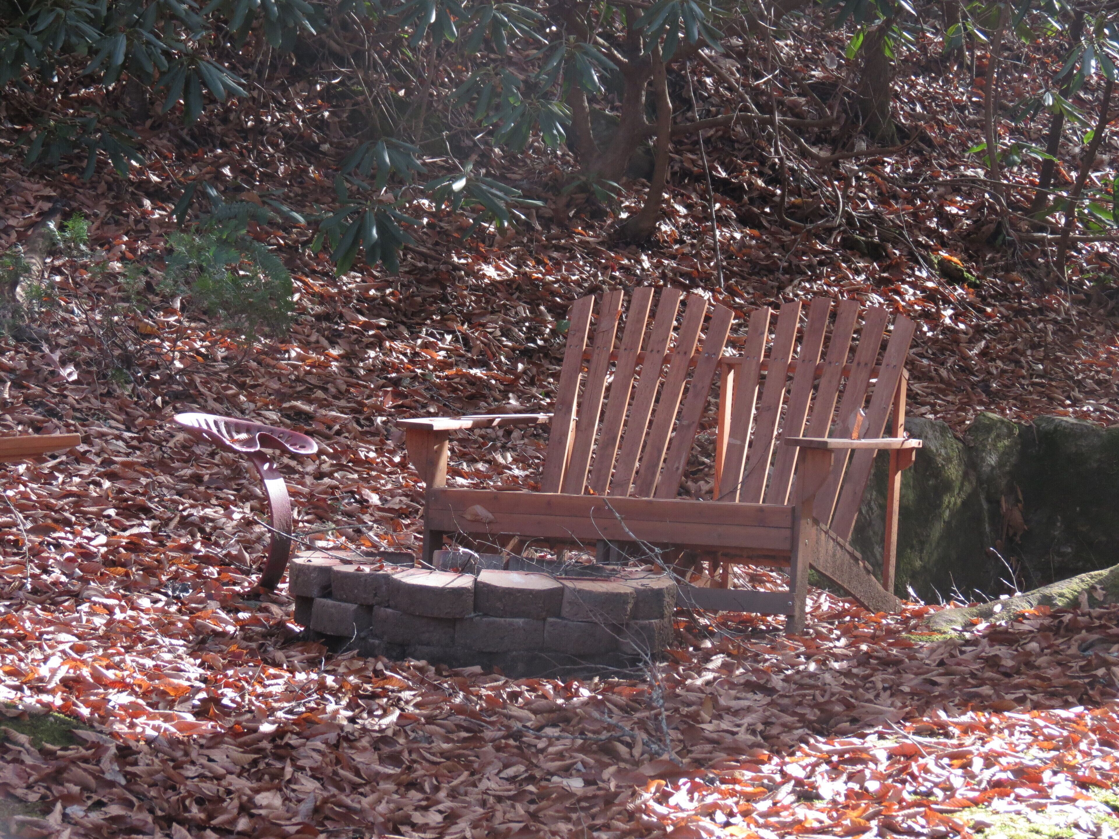 A wooden Adirondack chair with a stone fire pit in a forested area.