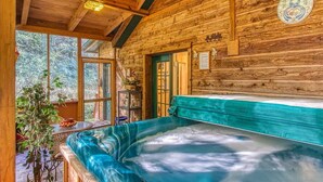 A wooden cabin with a hot tub, large windows, and a view of the forest.
