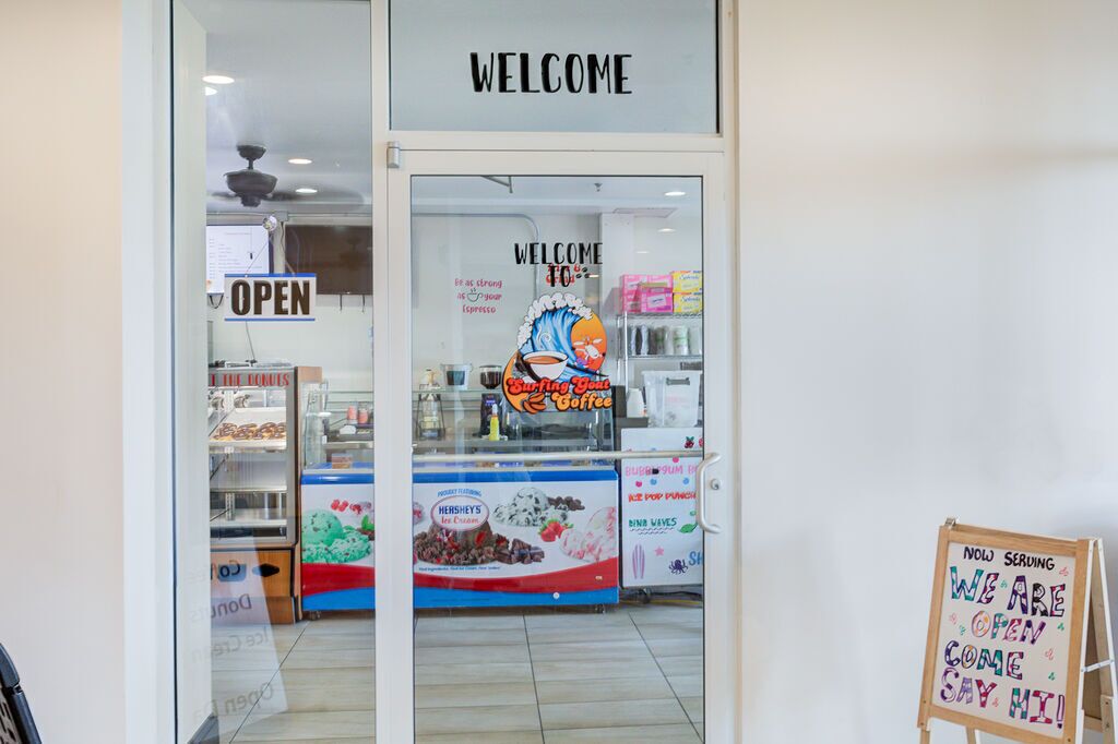 A coffee shop entrance with a 'WELCOME' sign, an 'OPEN' sign, and a menu board advertising 'Surfing Your Coffee'.
