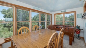 Una habitación con una mesa de madera, cuatro sillas, una mesa de fútbol de mesa y ventanas amplias con vistas a árboles.