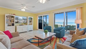 A spacious living room with a large sofa, a coffee table, and a bookshelf. There is a sliding glass door leading to a balcony with a view of the ocean. - Seagull-Gulf Front Home with Private Beach Crossover No Pets (Navarre)