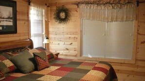 A cozy log cabin bedroom with a quilted bedspread, a wooden headboard, a wall-mounted clock, and a window with blinds. - Get Away from it All in This Immaculate Cabin in the Woods (Blairsville)