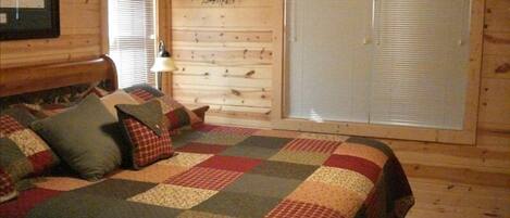 A cozy log cabin bedroom with a quilted bedspread, a wooden headboard, a wall-mounted clock, and a window with blinds.