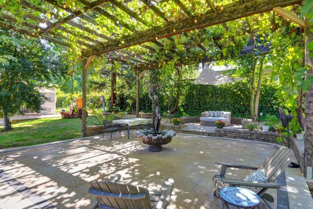 Een overdekt terras met een stenen fontein, zitplaatsen en een pergola met lichtslingers.
