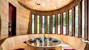 Une table ronde avec un plateau en verre, entourée de bancs en bois dotés de coussins rouges, est placée devant un mur de pierre agrémenté de grandes fenêtres donnant sur un paysage verdoyant.