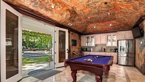 Une table de billard dans une pièce dotée d’un plafond en pierre, une cuisine équipée d’appareils électroménagers en acier inoxydable et une vue sur un jardin, visible à travers des portes vitrées.