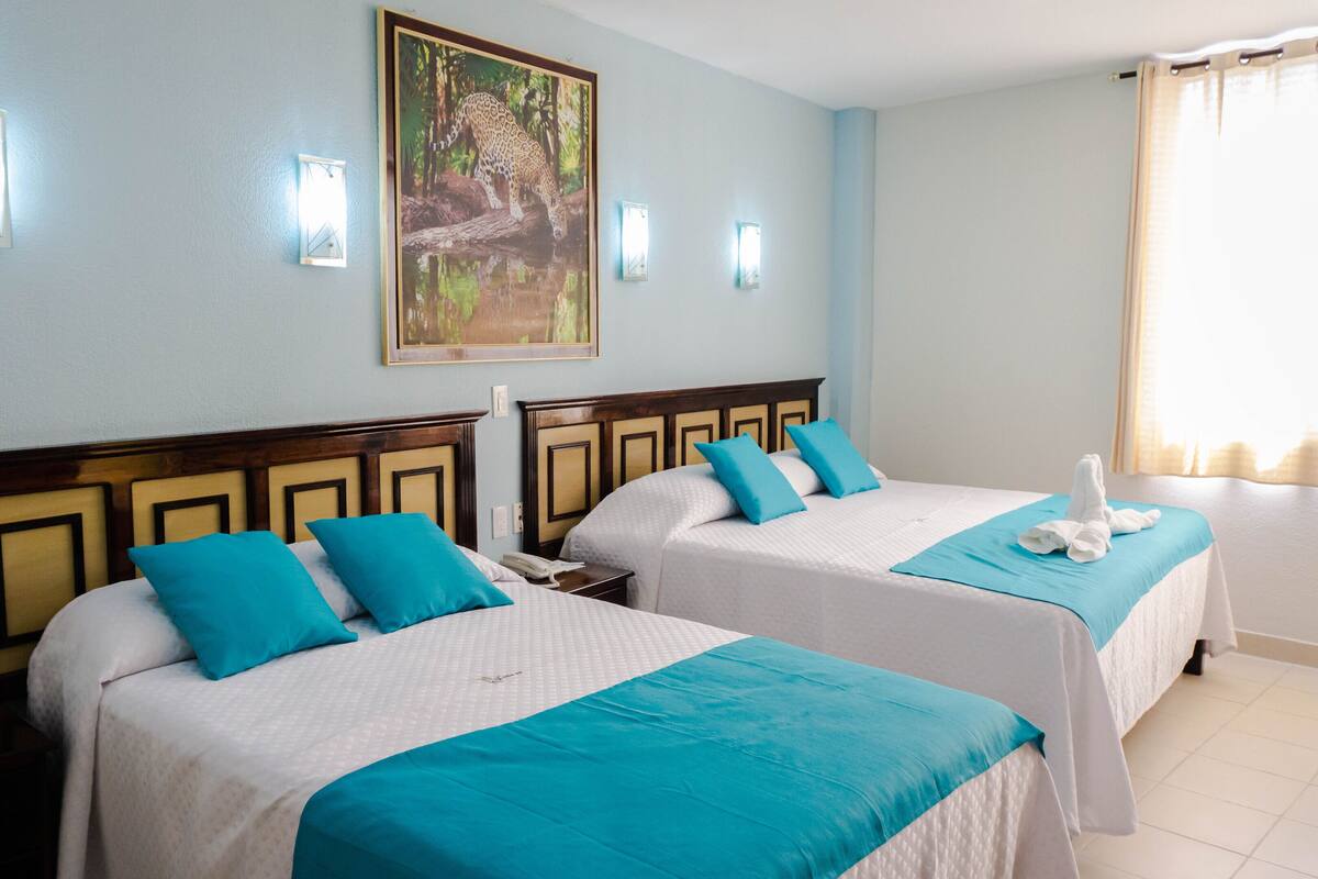 A hotel room with two beds, each with a wooden headboard and a blue accent pillow. There is a painting of a leopard on the wall above the beds.