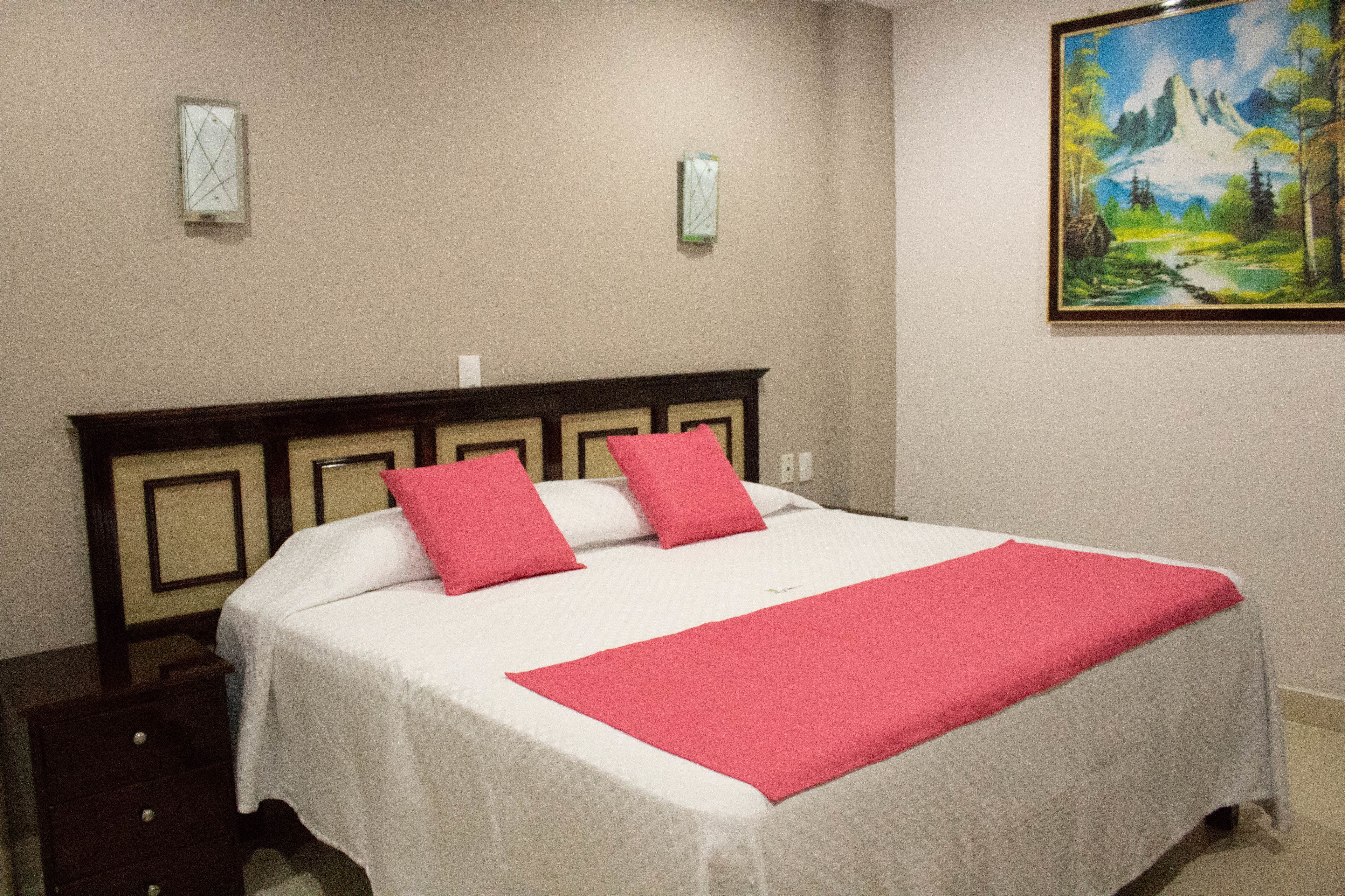 Una cama bien hecha con una colcha blanca y rosa, un cabecero de madera y un cuadro de un paisaje montañoso en la pared.