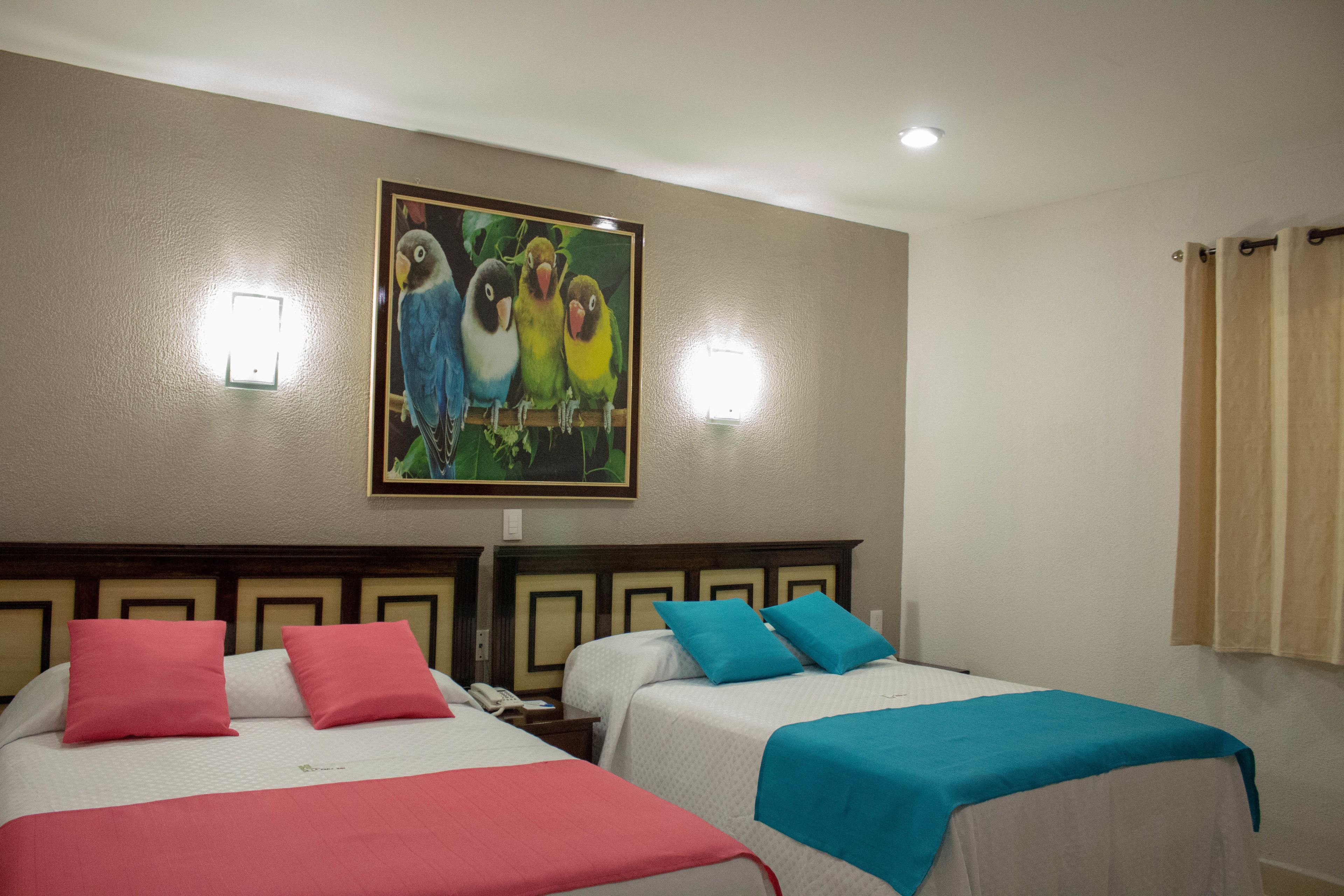 A hotel room with two beds, each with a different colored bedspread, a painting of birds on the wall, and a window with curtains.