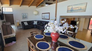 A modern living room with a black leather sofa, a dining table with blue placemats, and a red vase with white flowers.