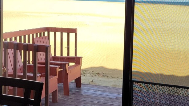 A room with a large window overlooking a sandy beach. There is a wooden table and chairs inside the room.