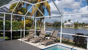 A covered pool area with lounge chairs, a clear view of a lake, and palm trees in the background. - Crown Pointe Home with Lake Views! (Naples)