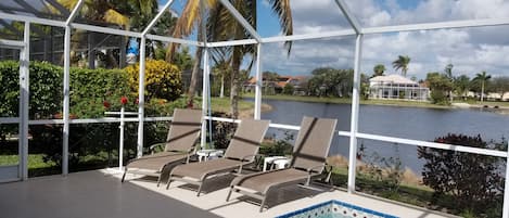 Un área de piscina techada con sillones de descanso, vista despejada de un lago y palmeras al fondo.