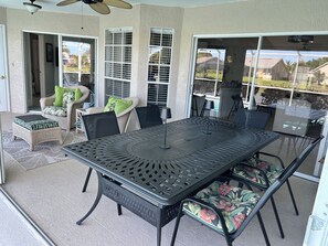 A covered patio area with a metal table and chairs, a wicker chair with cushions, and a view of the neighborhood through large windows. - Crown Pointe Home with Lake Views! (Naples)