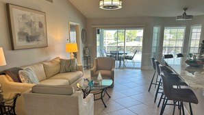 A living room with a sofa, coffee table, and chairs, a dining area with a table and chairs, and large windows with blinds. - Crown Pointe Home with Lake Views! (Naples)