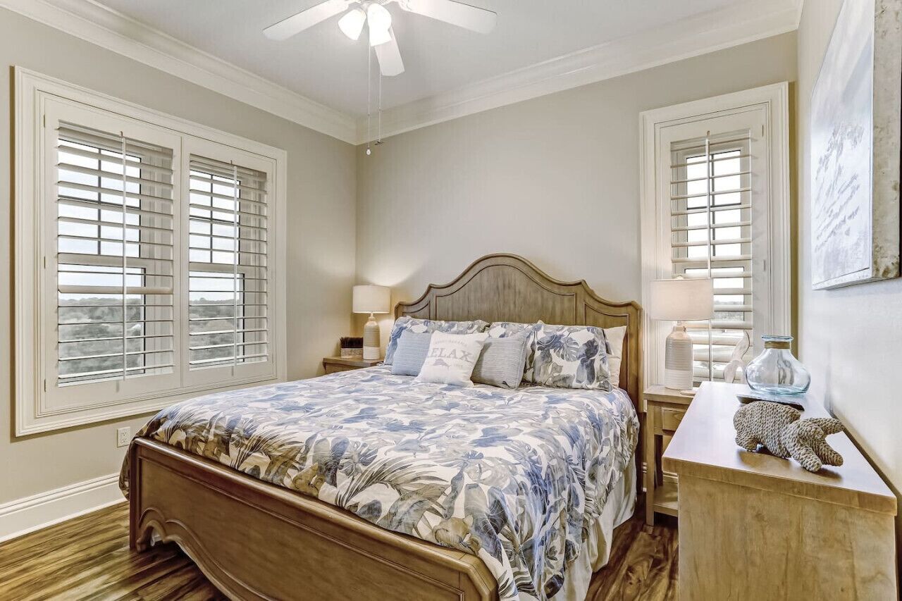 A bedroom with a wooden bed, two bedside tables, a ceiling fan, and windows with blinds.
