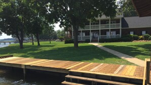 A wooden dock leading to a building with a balcony, surrounded by a grassy area and trees.