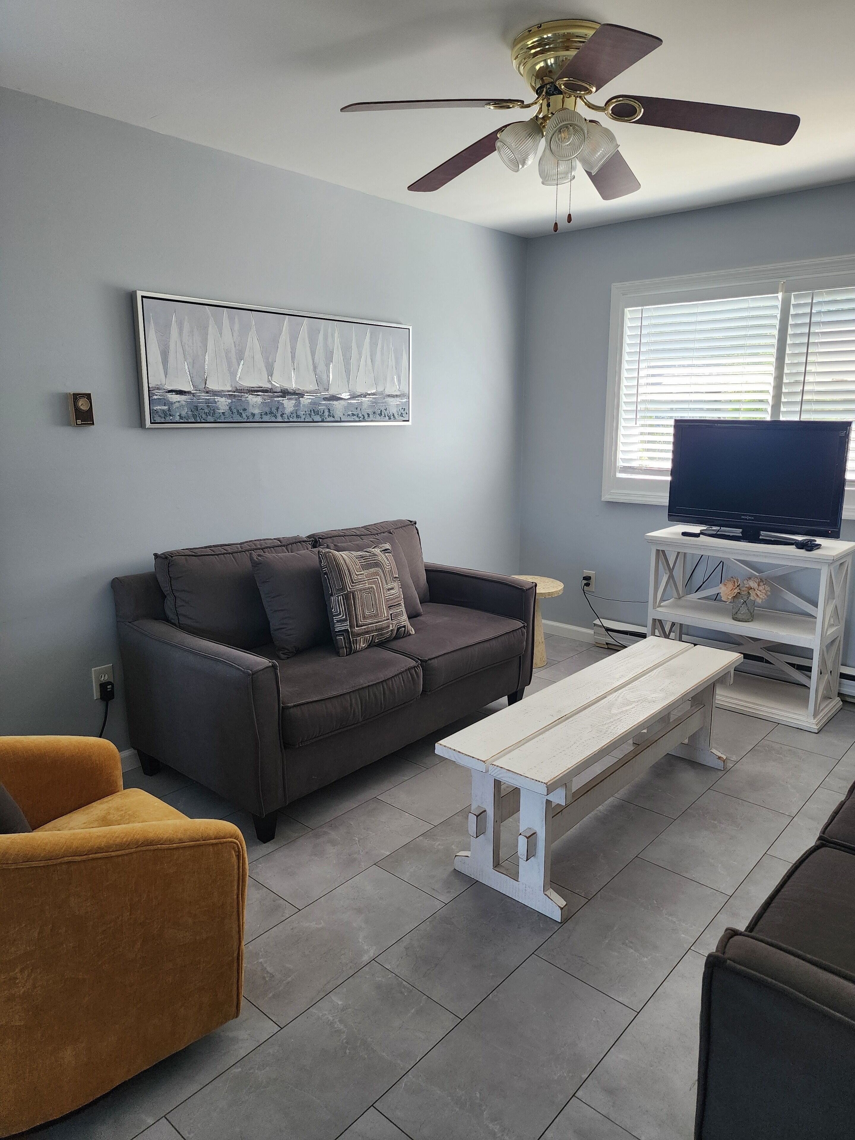 Un salon avec un canapé gris, un fauteuil marron, une table basse blanche, un ventilateur de plafond, un téléviseur posé sur un meuble blanc et une fenêtre avec des stores.
