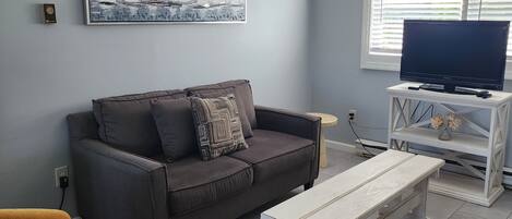 Un salon avec un canapé gris, un fauteuil marron, une table basse blanche, un ventilateur de plafond, un téléviseur posé sur un meuble blanc et une fenêtre avec des stores.