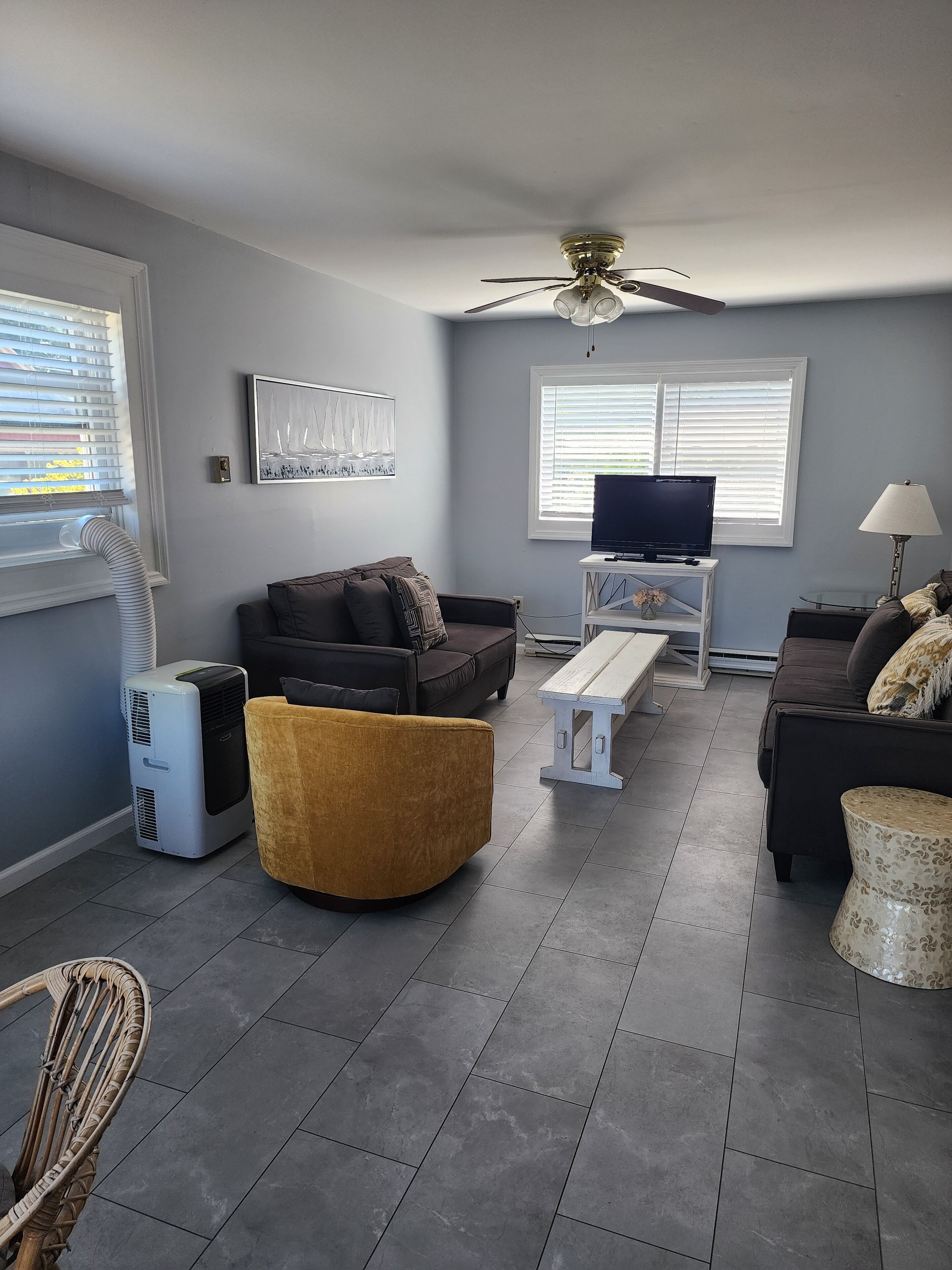 Un salon avec un canapé, un pouf jaune, un téléviseur posé sur un meuble et un ventilateur de plafond.