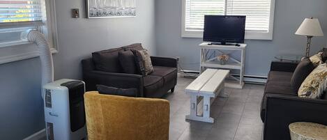Un salon avec un canapé, un pouf jaune, un téléviseur posé sur un meuble et un ventilateur de plafond.