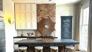 A kitchen with a central island, three wicker bar stools, a brick wall, and a menu board.