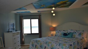 A bedroom with a bed, a dresser, a chair, a window with curtains, and a ceiling mural. - Sunglow, Beachfront Deluxe - 2 bed, 2 bath - ocean front unit (Daytona Beach Shores)