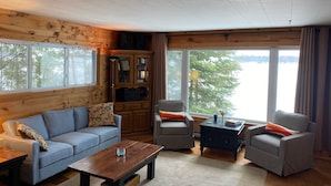 A cozy living room with a wooden interior, a sofa, two armchairs, a coffee table, and a cabinet with glass doors. - Green Lake: Charming Cottage with Sandy Beach & Dock - Ideal for Family Getaways (Interlochen)