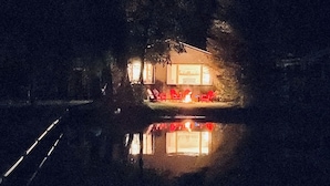 A nighttime view of a house with a lit interior, reflected in a body of water. - Green Lake: Charming Cottage with Sandy Beach & Dock - Ideal for Family Getaways (Interlochen)