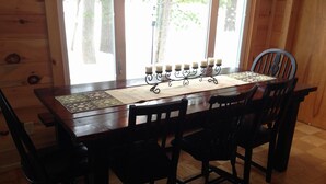 A wooden dining table with six chairs, a candle holder with candles, and a window with curtains. - Green Lake: Charming Cottage with Sandy Beach & Dock - Ideal for Family Getaways (Interlochen)