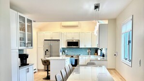 A modern kitchen with white cabinetry, a central island, and a dining area with a long table and chairs. - 1.5 blocks to Scarborough beach, Narragansett, fenced in yard, AC - New Updates! (Narragansett)
