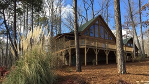 Eine Holzhütte mit einer Holzterrasse und großen Fenstern, umgeben von Bäumen und einem mit Laub bedeckten Boden.