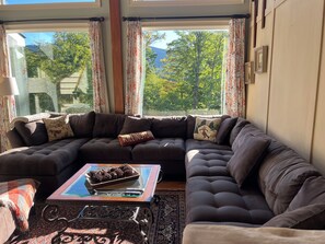 A modern living room with a sectional sofa, a glass coffee table with pine cones, and a view of a forest through large windows.