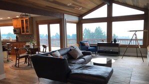 A spacious living room with a black leather sofa, a dining area with wooden furniture, and large windows offering a view of the outdoors. - Wine country with views (Newberg)