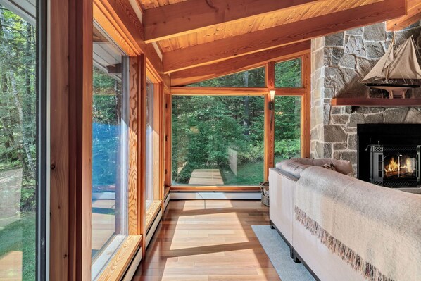 A cozy interior with a stone fireplace, wooden beams, and large windows offering a view of the outdoors. - Quechee House with Hot Tub, Sauna & Club Access (Quechee)