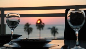Een tafel met twee wijnglazen en een schaal, met uitzicht op een zonsondergang en palmbomen.