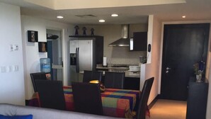 Een moderne woonkamer met een grijze bank, kleurrijke kussens, een eethoek met een tafel en stoelen, en een keuken met een koelkast en fornuis.