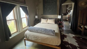 Une chambre avec un grand lit, un bureau et une chaise. Il y a une fenêtre avec des rideaux, un tableau noir accroché au mur et un tapis en peau de vache sur le sol.