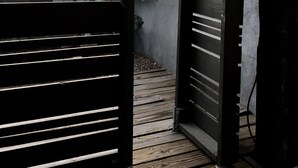 Une porte en bois, composée de lattes horizontales, qui donne sur un chemin en béton et un petit espace jardin agrémenté d’un arbre et de plantes.
