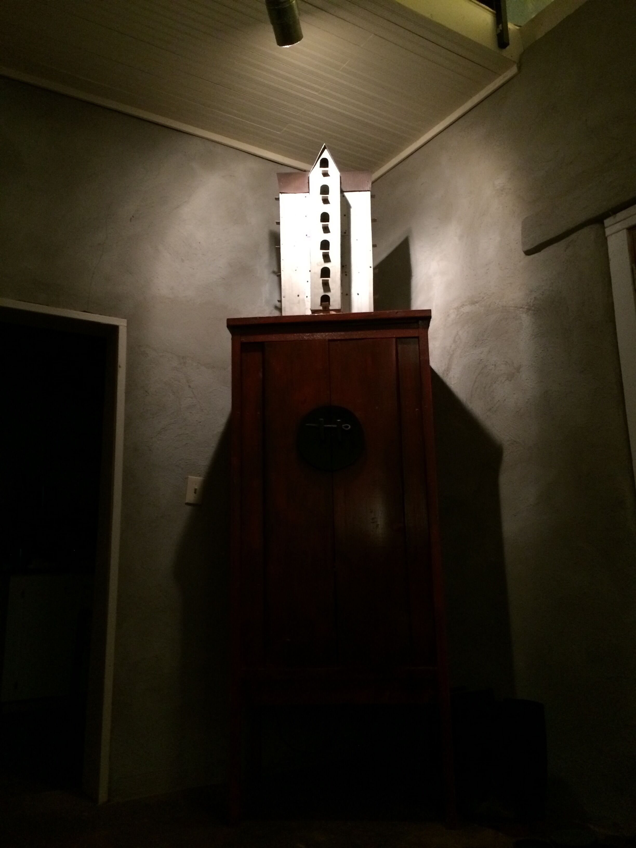 Un mueble de madera con un reloj y un objeto blanco encima, posiblemente decorativo, en una habitación con poca luz, con una puerta a la izquierda y una pared a la derecha.