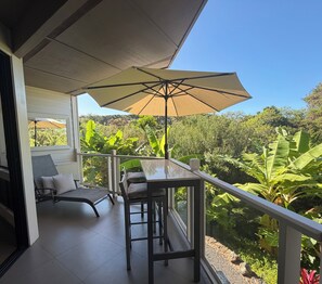 A balcony with a table and chairs, a large umbrella, and a view of lush greenery. - Luxury Wailea Villa l Stunning Remodel l 
Couples Retreat l Only 5 Star Reviews (Kihei)
