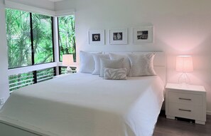 Ein Schlafzimmer mit einem großen Bett, weißer Bettwäsche, einem Nachttisch mit Lampe und einem Fenster mit Blick auf Grünflächen.
