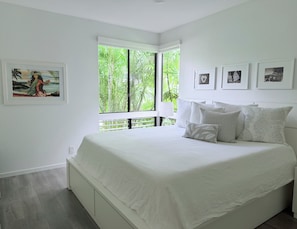 A bedroom with a large bed, a window with a view of greenery, and framed pictures on the wall. - Immaculate and Chic - Come relax in our luxury villa… (Kihei)