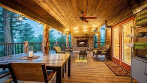 Une terrasse en bois avec une cheminée en pierre, des meubles en osier et une vue sur les arbres.