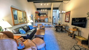 A spacious living room with a leather sofa, a coffee table, and a dining area with a wooden table and chairs. A kitchen is visible in the background, and a TV is mounted on the wall. - Luxury Condo w/ FREE WiFi, Parking, Heated Pool, Hot Tubs, Skier Shuttles (Avon)