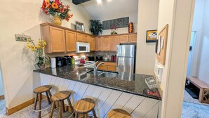 A kitchen with wooden cabinets, a stainless steel refrigerator, a microwave, and a granite countertop. There are three wooden stools and a potted plant. - Luxury Condo w/ FREE WiFi, Parking, Heated Pool, Hot Tubs, Skier Shuttles (Avon)