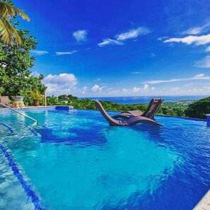 A pool with lounge chairs, clear blue water, and a view of the ocean and greenery. - Eagle`s Base Villa with Spectacular Views-Sleeps22 (Patience Hill)