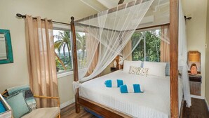 Ein Bett mit Baldachin, Holzboden, ein Sessel und Blick auf Palmen durch das Fenster.
