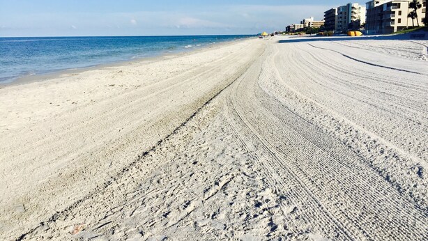 Une vaste plage de sable fin, une mer d’un bleu limpide et des bâtiments visibles au loin.
