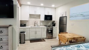 Une cuisine moderne avec des meubles blancs, un four à micro-ondes, une cuisinière et un réfrigérateur. On y trouve une table en bois et un lit recouvert d’une courtepointe à motifs floraux bleus.